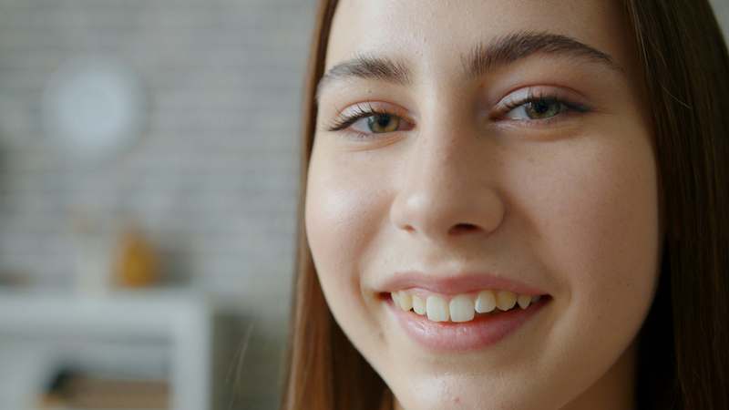 Woman with wavy brown hair smiling while applying a homemade aloe-vera gel to her cheek, natural lighting, kitchen counter background